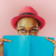 woman in red hat holds blue book over face